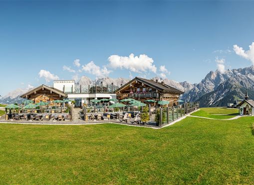 Eine schöne alpine Landschaft mit einem modernen Restaurant und traditionellen Chalets. Im Hintergrund sind majestätische Berge und ein klarer blauer Himmel zu sehen.