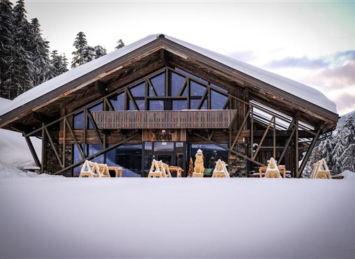 Ein modernes Holzhaus steht in einer verschneiten Landschaft. Im Hintergrund sind Bäume und ein klarer Himmel zu sehen.