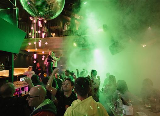 Eine lebendige Party in einem Nachtclub mit bunten Lichtern und Nebel. Leute tanzen und feiern unter einem großen Discokugel.