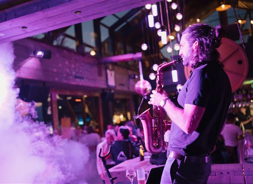 Ein Saxophonist spielt in einem stilvollen Veranstaltungsort mit bunten Lichtern und Rauch. Die Atmosphäre ist lebhaft und einladend.