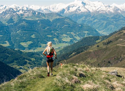 Eine Frau joggt auf einem Hügel mit einer atemberaubenden Aussicht auf grüne Täler und schneebedeckte Berge. Die Landschaft ist klar und einladend.