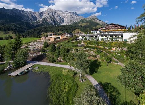 A picturesque landscape with a hotel, surrounded by mountains and a tranquil pond. The green surroundings are lush and inviting.