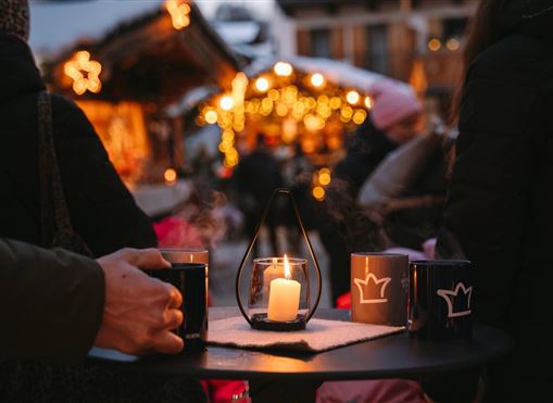 Eine festliche Szenerie mit Lichtern und einer Weihnachtsmarktbude. Auf einem Tisch stehen Getränke und eine Kerze, die eine gemütliche Atmosphäre schafft.