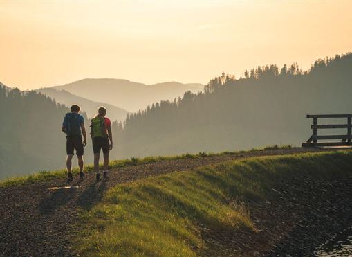 Zwei Wanderer gehen entlang eines Weges in einer bergigen Landschaft. Der Sonnenuntergang verleiht der Szene eine warme, goldene Atmosphäre.