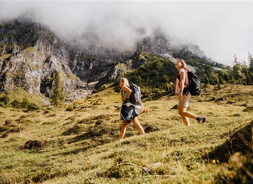 Zwei Wanderer erkunden eine malerische Berglandschaft. Die Umgebung ist grün mit Hügeln und Wolken über den Bergen.