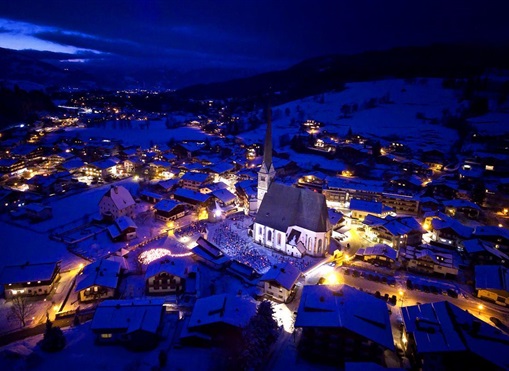 A picturesque winter landscape with snow-covered roofs and illuminated buildings. At the center stands a church, surrounded by a quiet, nighttime village.