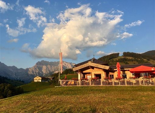 Eine gemütliche Berghütte mit Außenbereich und Sonnenschirmen. Im Hintergrund sind majestätische Berge und ein klarer Himmel zu sehen.