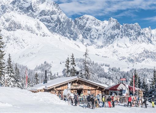 A picturesque mountain ski resort with a cozy hut in the foreground. In the background, majestic snow-covered mountains rise under a blue sky.