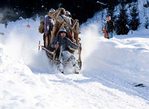 Eine Gruppe von Menschen fährt in einem selbstgebauten Schlitten durch den Schnee. Im Hintergrund sind verschneite Bäume und Zuschauer zu sehen.
